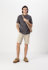 Camiseta gris con mangas enrolladas, pantalones cortos beige, zapatos marrones con suelas acanaladas y un bolso cruzado marrón. Diseño simple y colores neutros.
