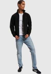 Sort lynlåsjakke med høj krave, båret over en hvid skjorte, parret med lyseblå jeans og sorte sneakers.