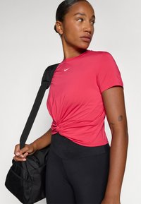 Červené športové tričko s krátkymi rukávmi, so uzlovým detailom vpredu, nosené s čiernymi legínami s vysokým pásom a čiernou športovou taškou.