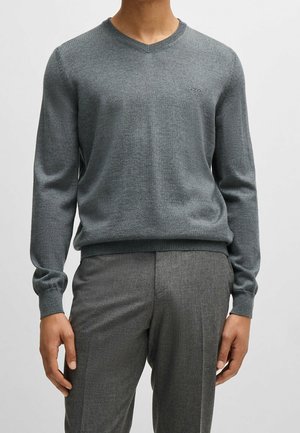 Mand iført tætsiddende grå V-hals trøje med lange ærmer og matchende grå skræddersyede bukser, stående foran en ensfarvet baggrund.