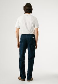 Camiseta blanca de manga corta y pantalones de pana azul oscuro, con bolsillos traseros visibles y un discreto detalle de logo en la cinturilla, mostrados desde atrás.