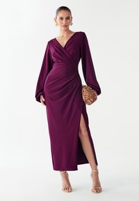 Lange pruimkleurige jurk met een V-hals, geplooide taille en wijde lange mouwen. Heeft een zijsplit en wordt gecombineerd met een gestructureerde gouden clutch.