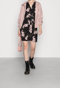 Abito nero floreale con cintura, abbinato a un giubbotto rosa chiaro con zip. Gli accessori includono una piccola borsa rosa e stivali neri con lacci.