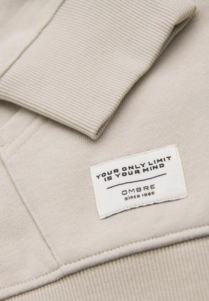 Lys beige sweatshirt med ribbet krave. Inkluderer et hvidt mærke med sort tekst, der står "DIN ENDETLIGE GRÆNSE ER DIN HJERNE" og "OMBRE siden 1985."