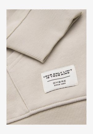 Jasno beżowy sweatshirt z prążkowanym kołnierzem. Zawiera białą metkę z czarnym napisem "TWÓJ JEDYNY LIMIT TO TWOJA MYŚL" oraz "OMBRE od 1985 roku."