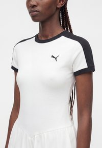 Femme portant une robe blanche avec un bord noir aux épaules et aux manches, avec un petit logo Puma noir sur la poitrine.