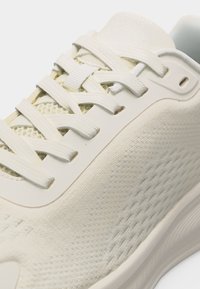 Gros plan sur une basket blanche avec un tissu texturé, des lacets plats et une languette rembourrée, mettant en valeur le design léger et respirant de la chaussure.