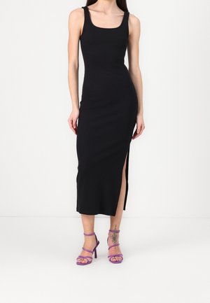 Femme portant une robe midi noire sans manches avec une fente latérale et des sandales à talons hauts violettes, montrant un tatouage sur sa jambe inférieure droite.