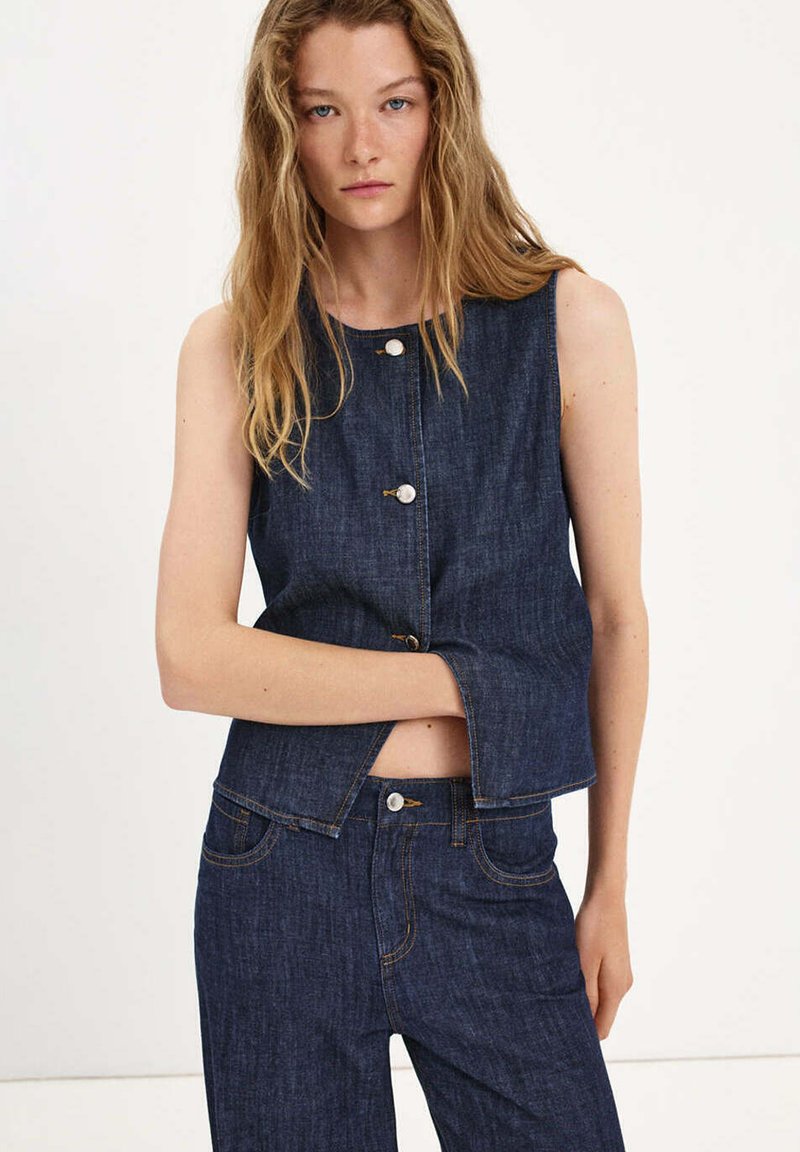 Young woman with long blonde hair wearing sleeveless dark denim vest and matching jeans against plain background.