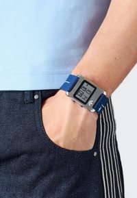 Digitális óra téglalap alakú ezüst házzal és kék szilikon pánttal, amely egy digitális kijelzőt tartalmaz, amely vastag számokkal mutatja az időt.