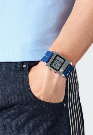 Digitalna ura s pravokotno srebrno ohišje in modrim silikonskim trakcem, ki ima digitalni zaslon, ki prikazuje čas v velikih številkah.
