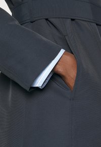Tumedast hall trench coat, millel on sile tekstuur, vöö, kaldenurgaga taskud ja käiste alt paistmas helesinine särgi mansett.
