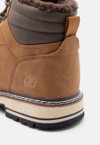 Botte en cuir brun avec un col rembourré gris, présentant une surface texturée, des coutures blanches et une semelle en caoutchouc pour une meilleure adhérence.