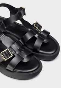 Sandalias de cuero negro con correas anchas, hebillas metálicas y suela gruesa. Textura suave y diseño minimalista con suela de goma.