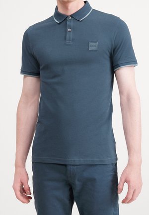 Mężczyzna ubrany w dopasowaną ciemnoniebieską koszulkę polo z subtelną naszywką z logo na lewej piersi oraz pasujące niebieskie spodnie.
