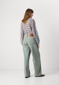 Lys grønn vide jeans med en brun lærlapp på baksiden. Modellen har på seg en gjennomsiktig rutete langermet topp. Svarte flate sko.