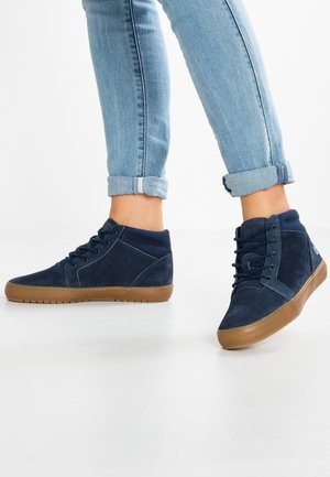 Person iført opsmøgede lyseblå jeans og marineblå høje sneakers med brune såler, stående på et hvidt gulv.