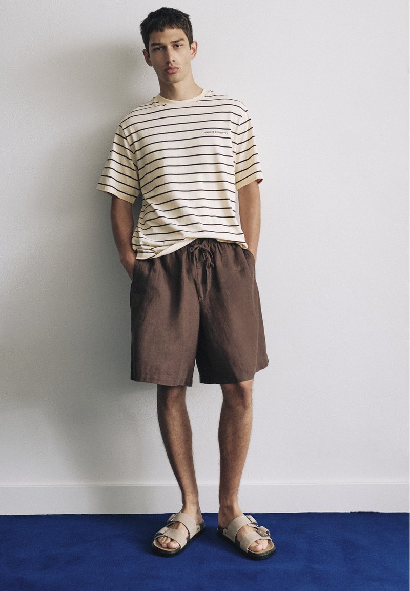 Jeune homme debout contre un mur portant un t-shirt beige rayé, un short marron et des sandales beiges ouvertes à l'avant sur un sol bleu. Mains dans les poches.