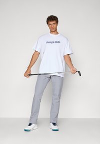 Hvit oversized t-skjorte med "Range Rats" grafikk, lysegrå bukser og hvite sko med blå detaljer, holder en golfklubbe.