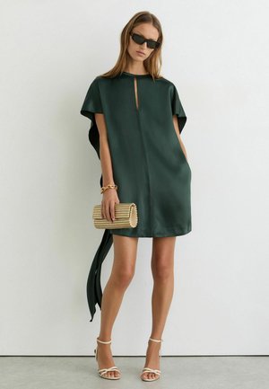 Vrouw draagt een donker groen kort jurkje met strikdetail aan de achterkant, houdt een beige clutch vast, draagt witte hakken met bandjes en een zwarte zonnebril, staat tegen een witte muur.