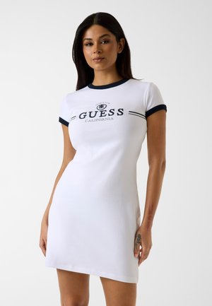 Mujer con vestido blanco de manga corta con ribete negro y el logo "GUESS California", posando frente a un fondo blanco liso.