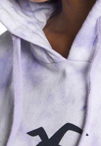 Fialová mikina s tie-dye motivem, která má klokaní kapsu a kapuci se stahovací šňůrkou. Na přední straně je výrazné tmavé logo. Vyrobena z měkké bavlny.