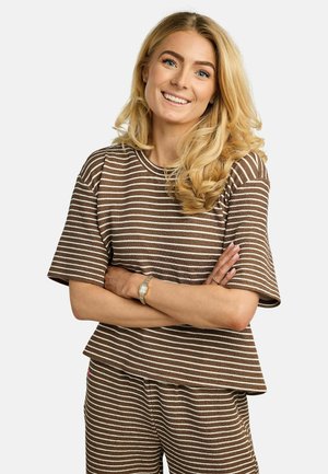 Femme souriante aux longs cheveux blonds, portant un haut à manches courtes rayé marron et blanc ainsi qu'un pantalon assorti, les bras croisés, debout devant un fond blanc.