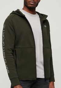 Felpa con zip verde oliva realizzata in tessuto morbido, dotata di cappuccio, tasche laterali, dettagli grigi e grande logo "SUPERDRY" sulla manica.