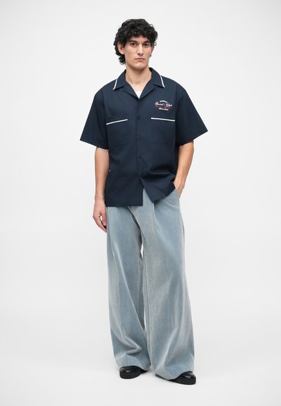 Chemise à manches courtes marine avec des accents blancs et de la broderie, assortie à un pantalon en denim clair à jambe large. La tenue présente une coupe décontractée.