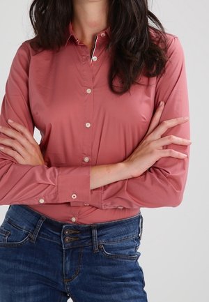 Kobieta o ciemnych włosach, ubrana w różową koszulę z długim rękawem zapinaną na guziki i niebieskie dżinsy, z założonymi na piersi ramionami.