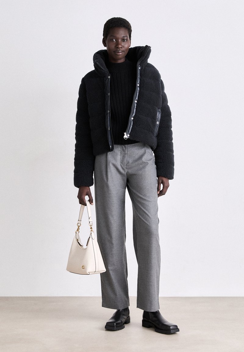 Giacca a vento nera con texture pelosa, maglione nero a coste, pantaloni grigi su misura e borsa bianca con dettagli dorati. Stivali neri.