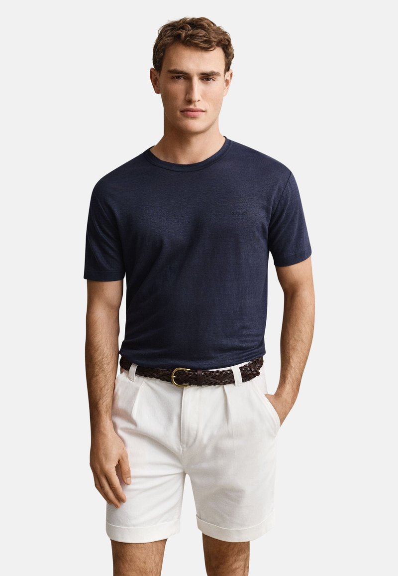 Homme portant une chemise bleu marine à manches courtes, un short blanc avec une ceinture tressée marron, debout devant un fond uni.