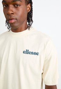 Majica od pamuka boje krem sa okruglim izrezom i kratkim rukavima. Na prsima se nalazi podignuti tamnoplavi logo "ellesse".