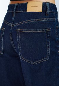 Tmavomodré denimové džínsy s oranžovým prešívaním, s koženou nálepkou s nápisom „PULL&BEAR“ na páse a klasickými zadnými vreckami.