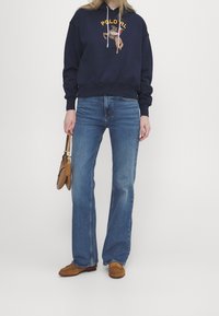 Felpa con cappuccio blu navy con emblema; jeans azzurri a zampa; scarpe in pelle scamosciata marrone; porta una piccola borsa marrone chiaro. Colori nitidi e design casual.