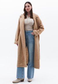 Beige wollen oversized jas met brede revers, gecombineerd met een licht tan aangesloten top en hooggeplaatste spijkerbroek met uitlopende pijpen in blauw. Lichte enkellaarzen.