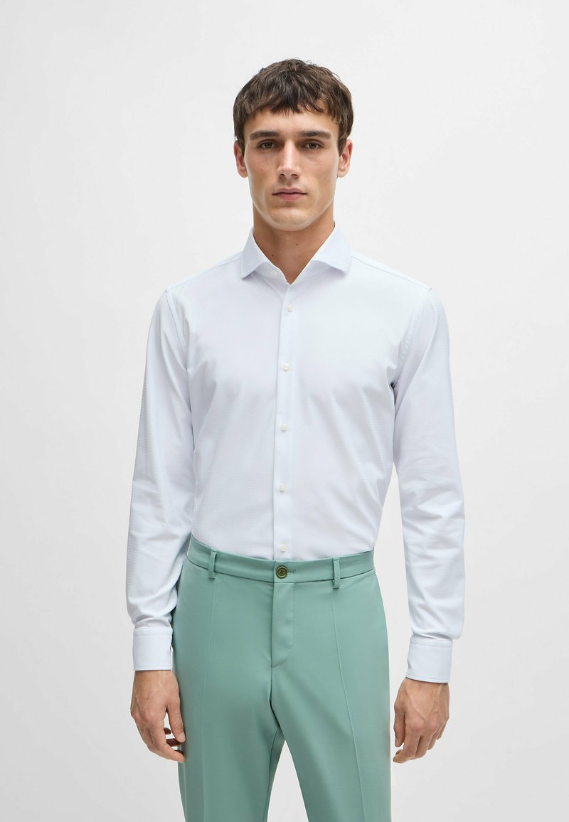 Chemise blanche à manches longues avec une finition texturée et un col à boutons, associée à un pantalon tailleur vert menthe et un détail de bouton distinct.