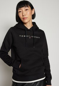 Sudadera negra de tejido suave con un bolsillo canguro frontal, capucha ajustable con cordón, y el logo "TOMMY JEANS" en el pecho.