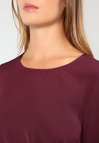 Burgundikjol med rund halsringning och slät textur, med en subtil lyster och utan synliga mönster eller detaljer.