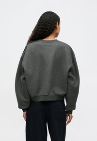 Grauer übergroßer Sweatshirt mit Rundhalsausschnitt, gerippten Bündchen und Saum. Weiches, strukturiertes Material mit abgesenkten Schultern, kombiniert mit dunklen Jeans.