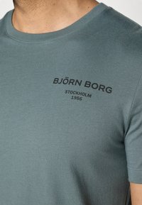 T-shirt teal a maniche corte realizzato in cotone, con collo a giro e testo stampato in nero "BJÖRN BORG STOCKHOLM 1956" sul davanti.