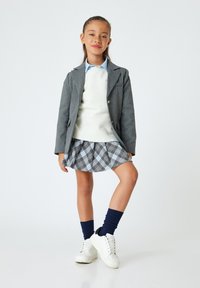 Blazer grigio, maglione bianco, camicia azzurra con colletto, gonna a quadri grigia e blu, calze blu navy e sneakers bianche con dettagli neri.