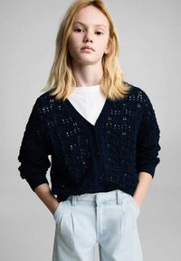 Cardigan in maglia navy con texture a traforo e chiusura a bottoni, abbinato a pantaloni alta vita azzurri chiari con pieghe.