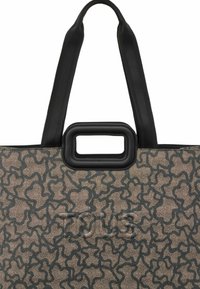 Torba na ramię z wzorzystym materiałem w brązowo-czarnym kolorze, z prostokątnym czarnym uchwytem i wytłoczonym logo na środku. Miękka tekstura.