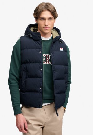 Young man wearing a dark blue padded vest with hood over a green sweatshirt and beige pants, standing with one hand in pocket.