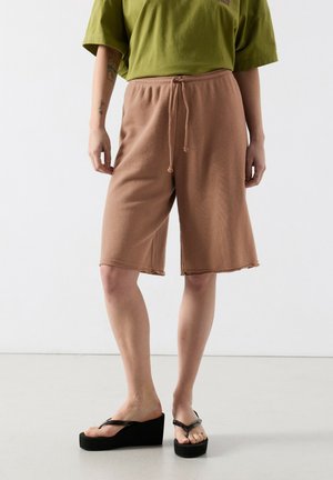 Person iført brune knælange shorts med snøre, grøn oversized t-shirt og sorte kilehøjhælede flip-flops, stående mod en ensfarvet hvid væg.