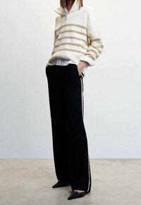 Gestreifter, weiß-beiger Strickpullover mit halbem Reißverschlusskragen, kombiniert mit schwarzen weitem Hosen mit Seitstreifen und schwarzen flachen Schuhen.