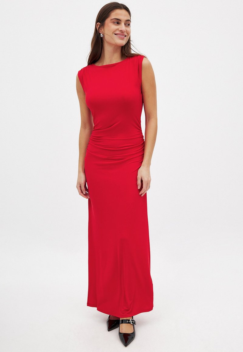 Czerwona, bezrękawna sukienka maxi o gładkiej fakturze i marszczonym boku. Połączona z czarnymi, szpiczastymi butami.