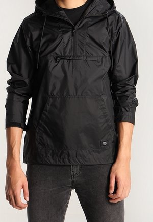 Osoba ubrana w czarną wiatrówkę z kapturem, z przednią kieszenią na zamek i kangurzą kieszenią, w połączeniu z ciemnoszarymi dżinsami.