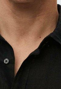 Camisa negra de botones con una textura suave, que presenta un cuello redondeado y un detalle de botón cerca del cuello, mostrando una vista en primer plano.